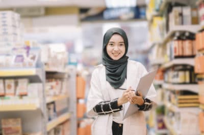 A female pharmacist wearing a hijab stands smiling and facing the camera while holding a clipboard and pen