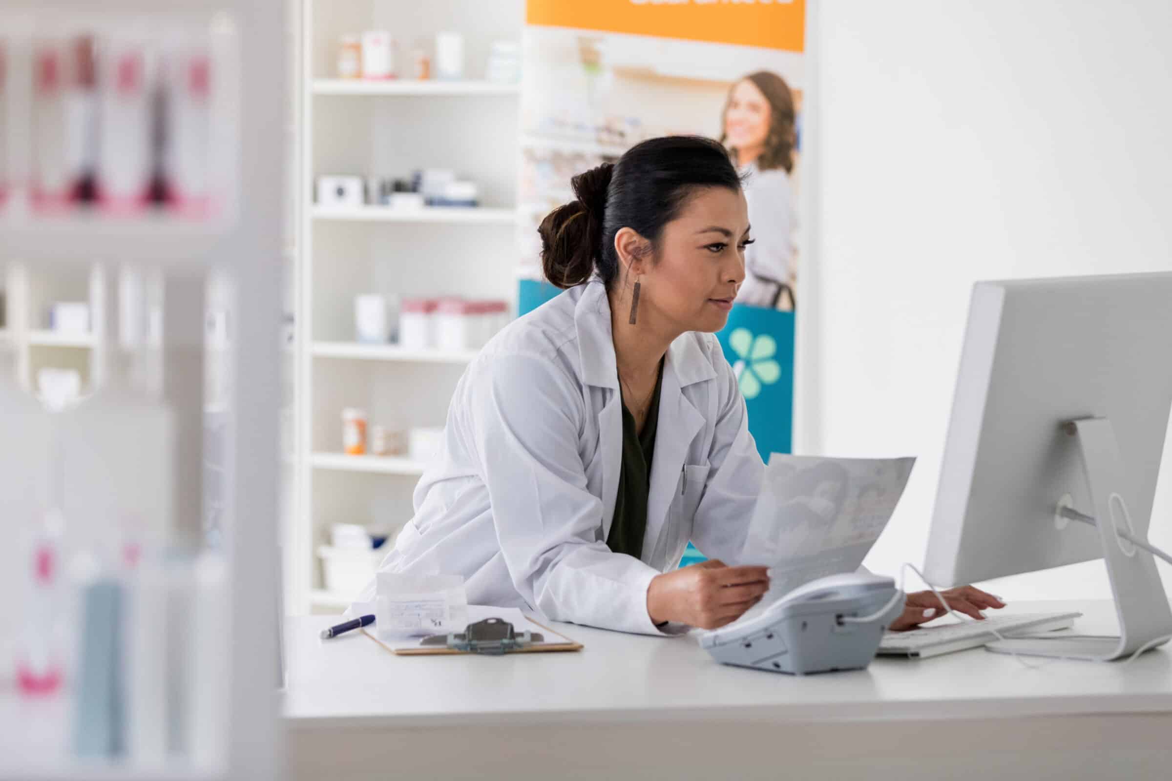 A pharmacist leaning on a pharmacy counter and typing on a computer.